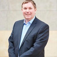 Portrait of Dr. Scott Hollier standing against a limestone wall, outside. Scott wears a blue suit and holds his arms in front of him.