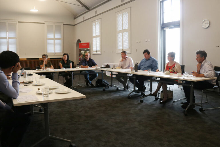 the board members sitting at long tables arranged in a C-shape.