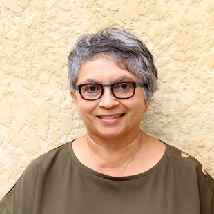 Jackie Weinman looking straight at the camera and smiling. She wears glasses and a olive coloured shirt with buttons on the left shoulder.