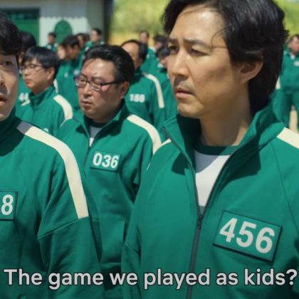 Image of group of men who are characters from Squid Game. They are wearing green and white tracksuits, all with numbers on their jackets. The one on the left who is numbered 118 is asking the one on the right numbered 456 which has been captioned, "The game we played as kids?" 