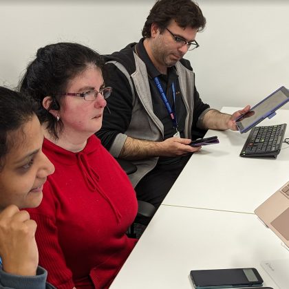 An image of three people, two women and a man, at their workstation, where they are working on a tablet and laptops. The woman closest to view is pointing to the headset she is wearing.