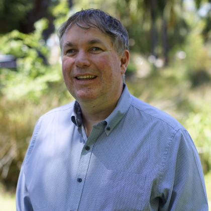 Photo of Scott Hollier, Man dressed in Blue shirt standing in front of trees smiling