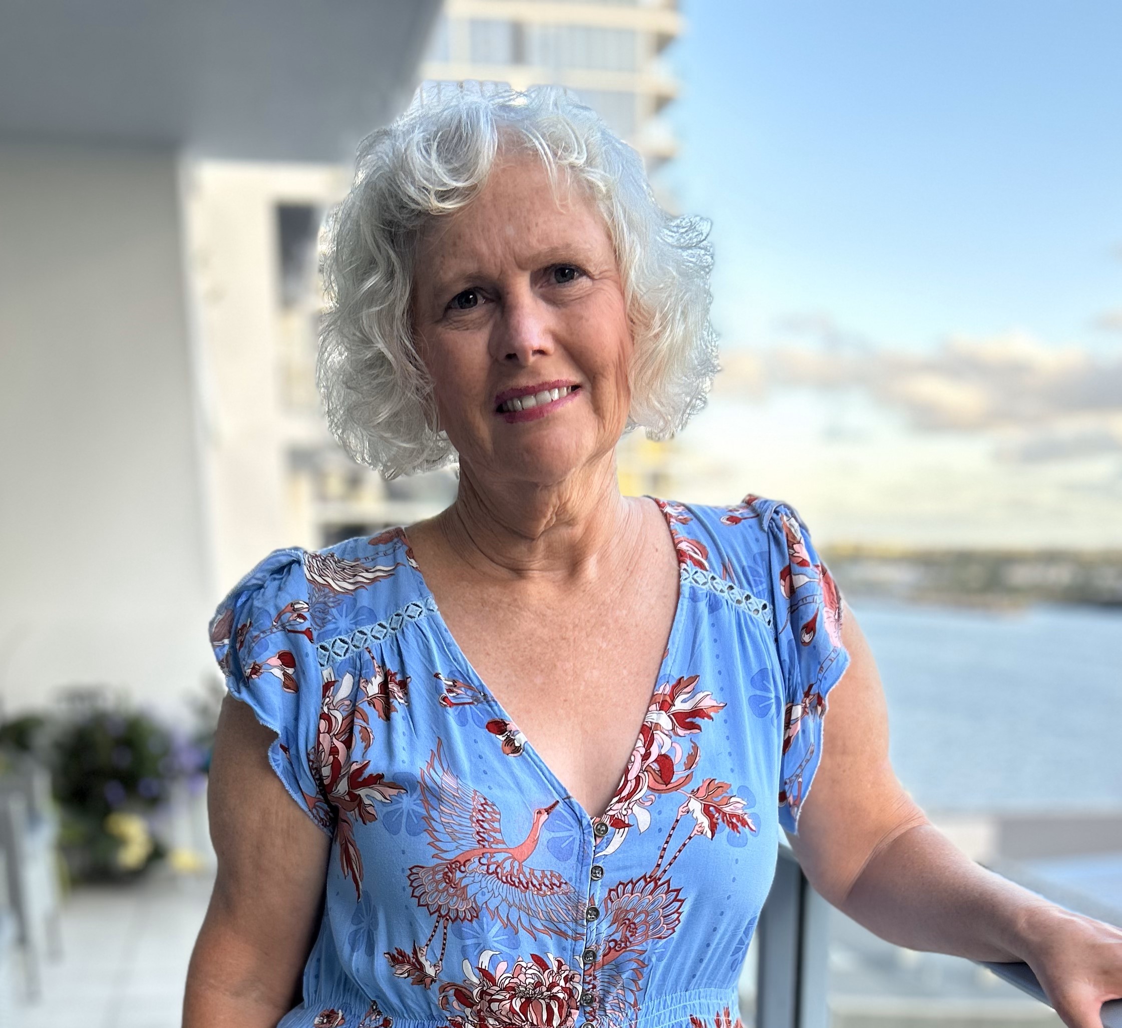 Narelle smiles at the camera, wearing a light blue dress with red and white floral patterns.