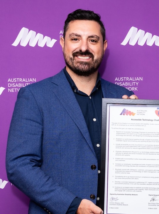 Ben, holding a framed certificate, smiles at the camera, with a purple background behind him.