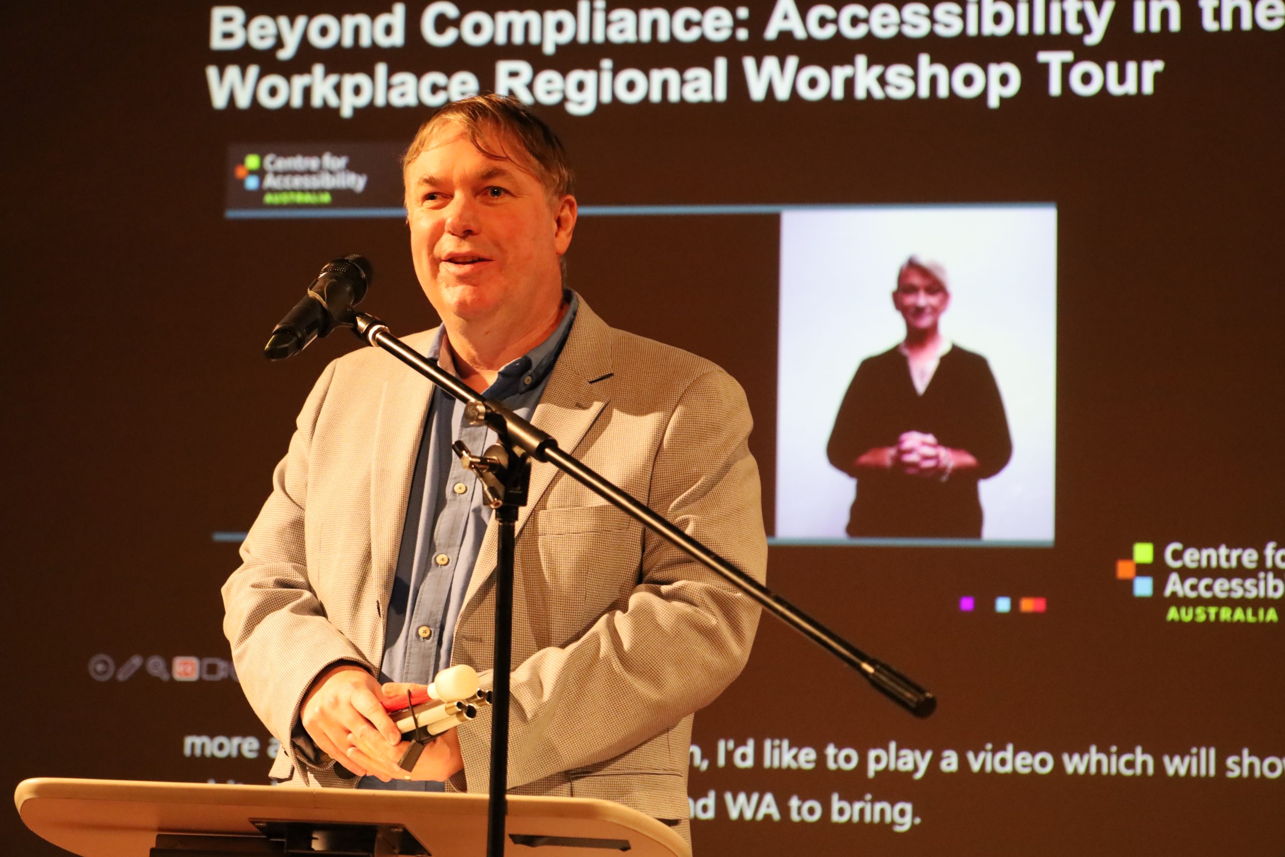 Dr. Scott Hollier stands, speaking into a microphone. Behind him is a screen with a Powerpoint presentation playing about the accessibility workshops.