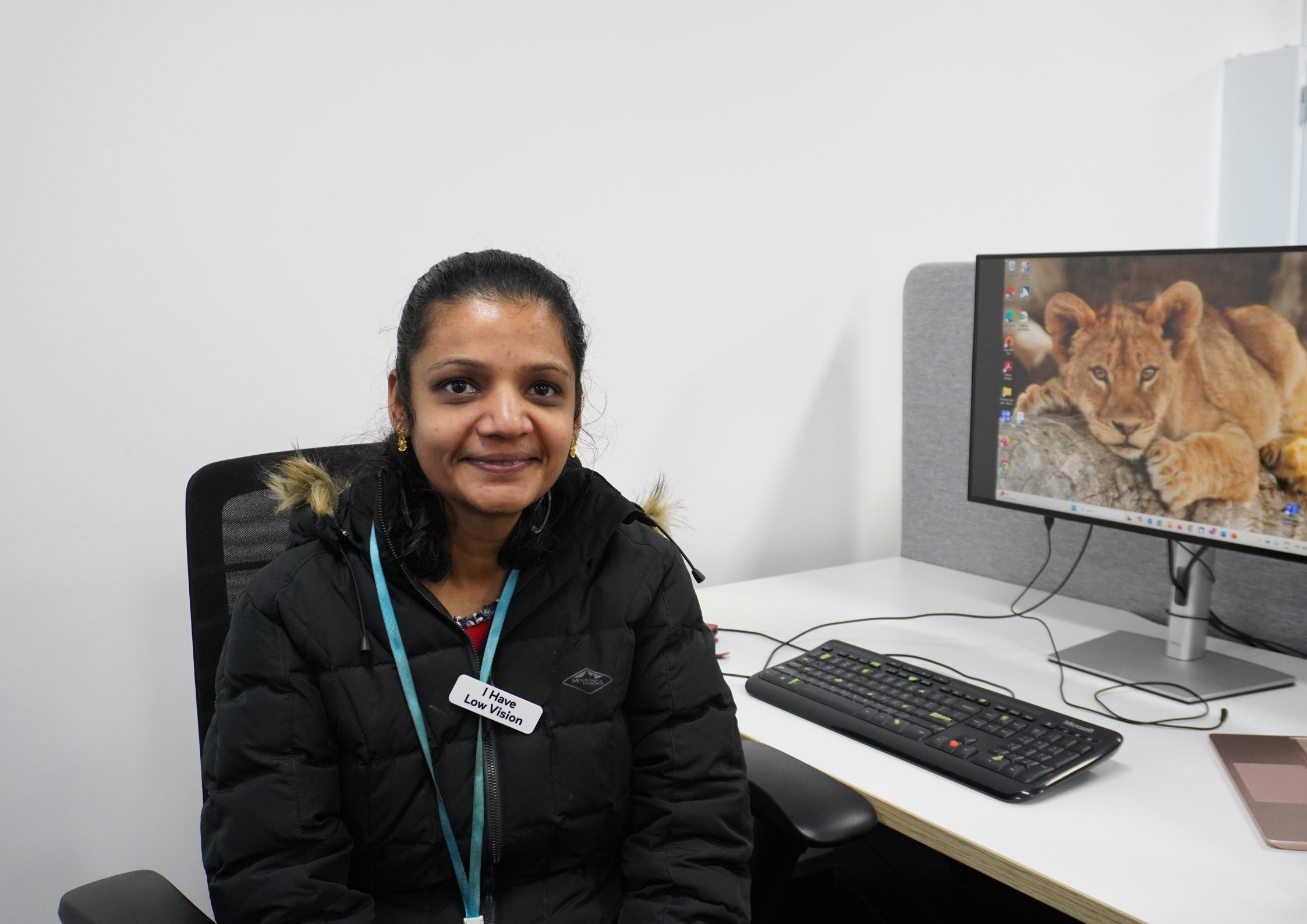 Aarthi is wearing a coat, with a fur lining. She is smiling, and sitting in front of a computer.