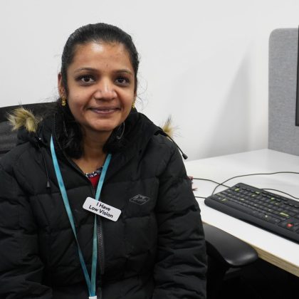 Aarthi is wearing a coat, with a fur lining. She is smiling, and sitting in front of a computer.