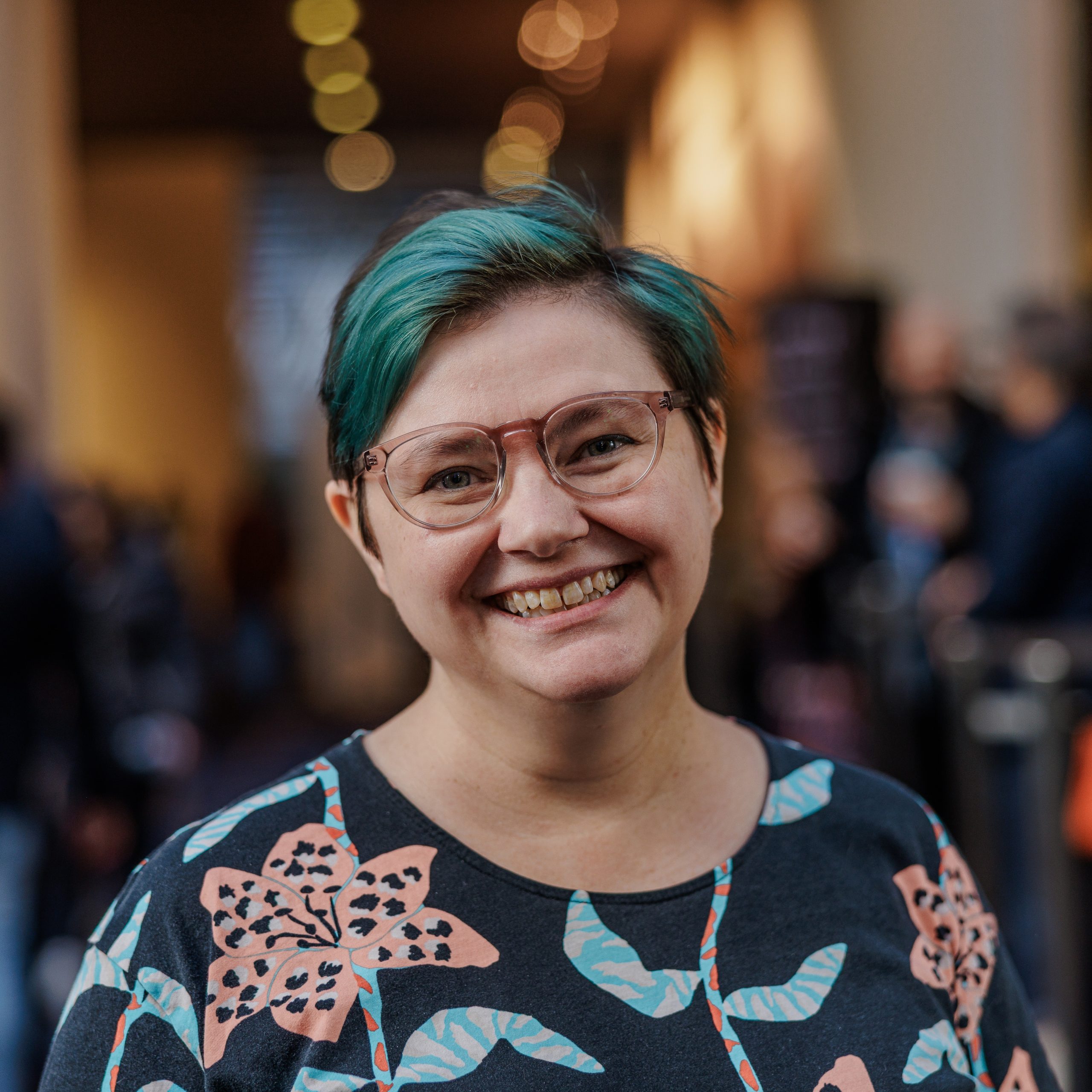 Julie Grundy smiles at the camera. She is wearing a dark blue shirt with a floral pattern. Her hair is dyed blue-green.