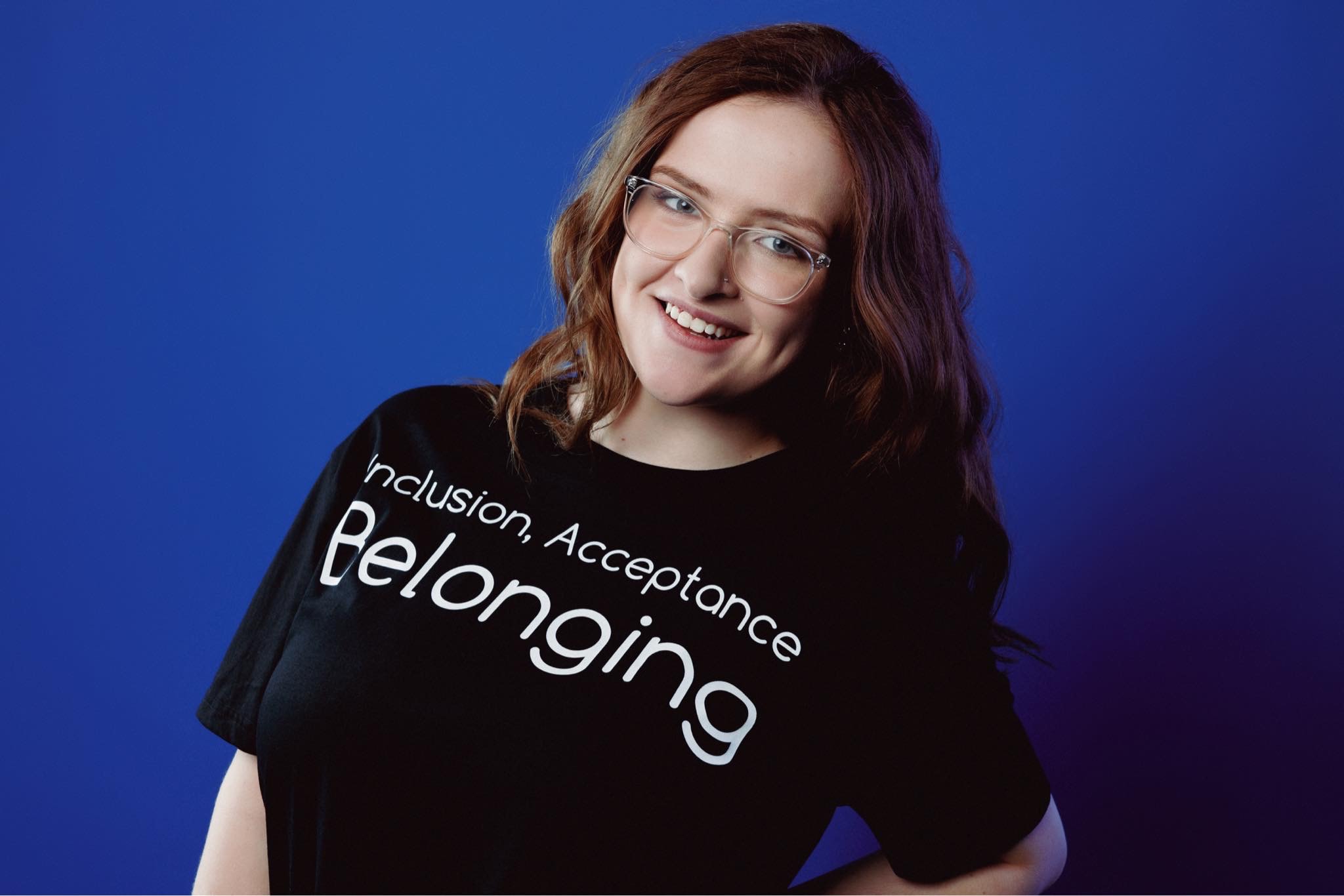 A young woman smiles at the camera. The background is blue. She wears a black shirt that says inclusion, acceptance, belong.