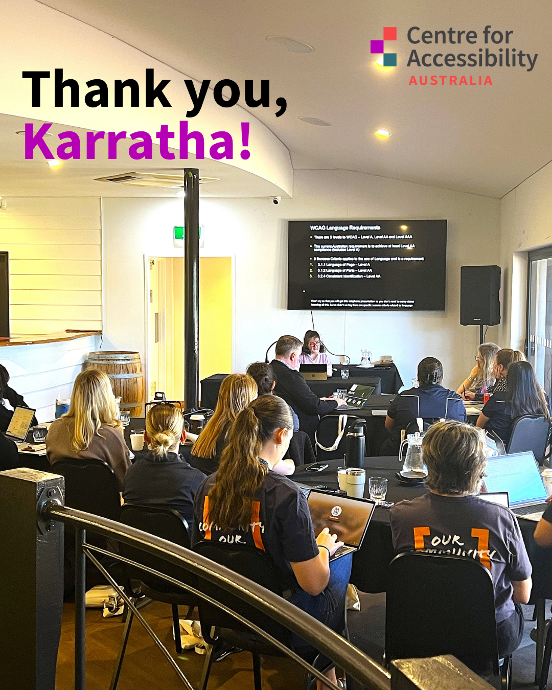 A group of people sit in a conference-style workshop in a bright meeting room, many working on laptops while facing a presentation screen at the front. Several attendees wear black shirts with “Tour Accessibility” branding on the back. Large text overlaid on the image reads, “Thank you, Karratha!” in black and purple lettering.
