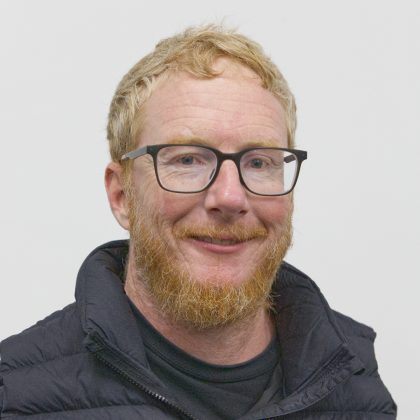 Close-up portrait of Matt. He has short ginger-blond hair, a ginger beard and black rectangular glasses. He is wearing a black puffer jacket against a plain white background.