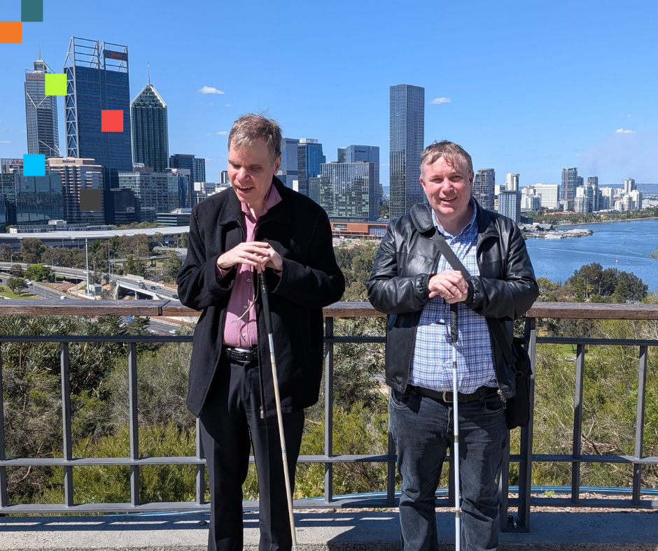 Image of Dr. Jason White and Dr Scott Hollier posing together for a photo. The background comprises of an outdoor environment, against a backdrop of tall buildings and a river.